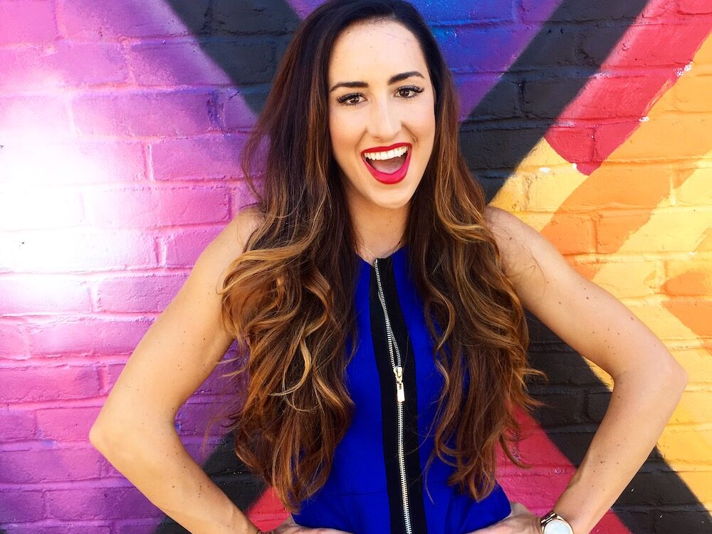 Codie Sanchez stands smiling and with her hands on her hips in front of a colorful geometric mural.