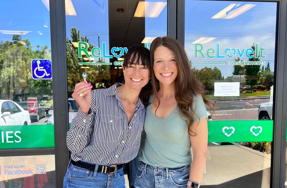 Two women pose, smiling, in front of a storefront with the name, ReLoveIt. One of the women is holding up a key.