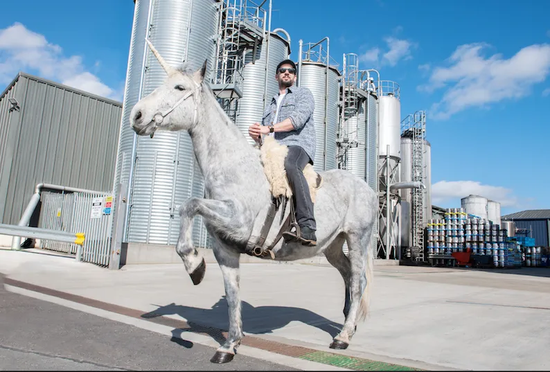 James Watt sits in a saddle atop a white and grey-speckled unicorn, standing outside a beer brewing facility.